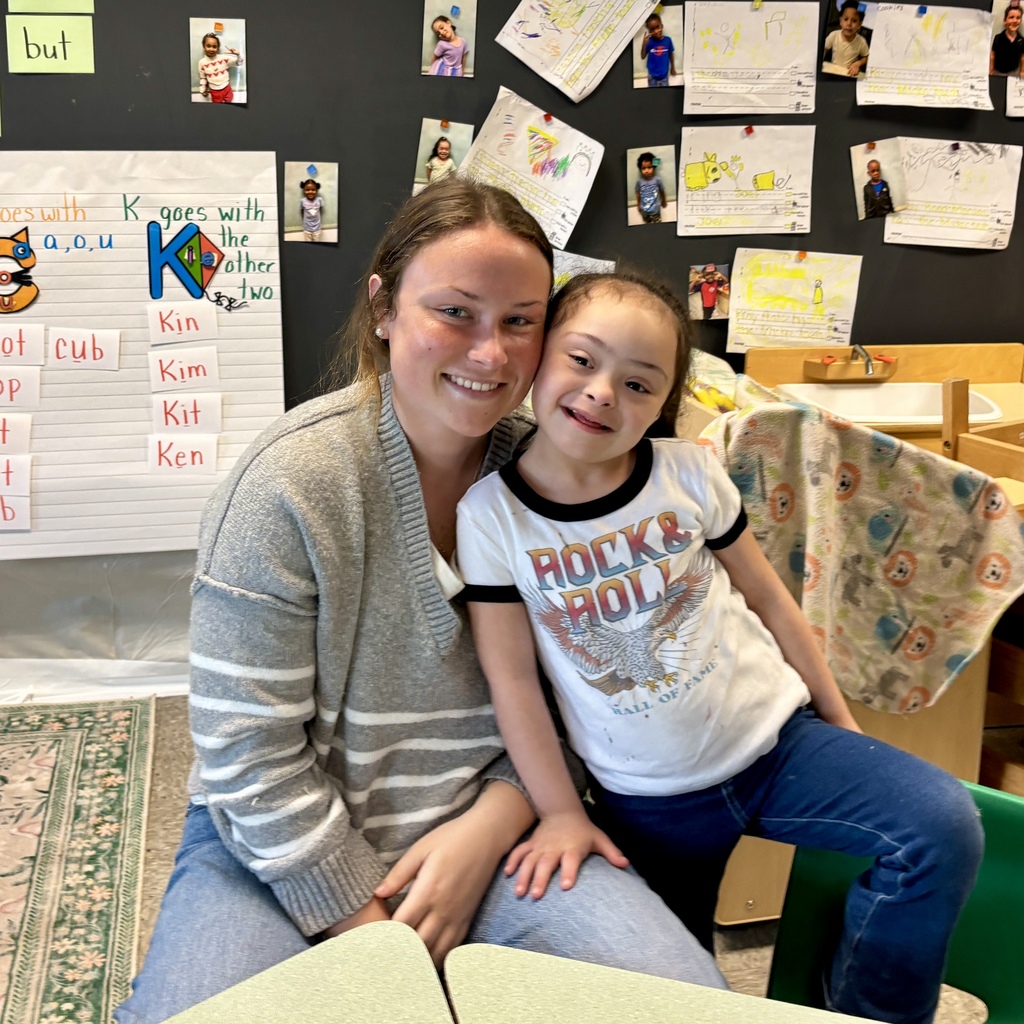 A smiling young woman and a girl with Down syndrome pose together in a classroom.