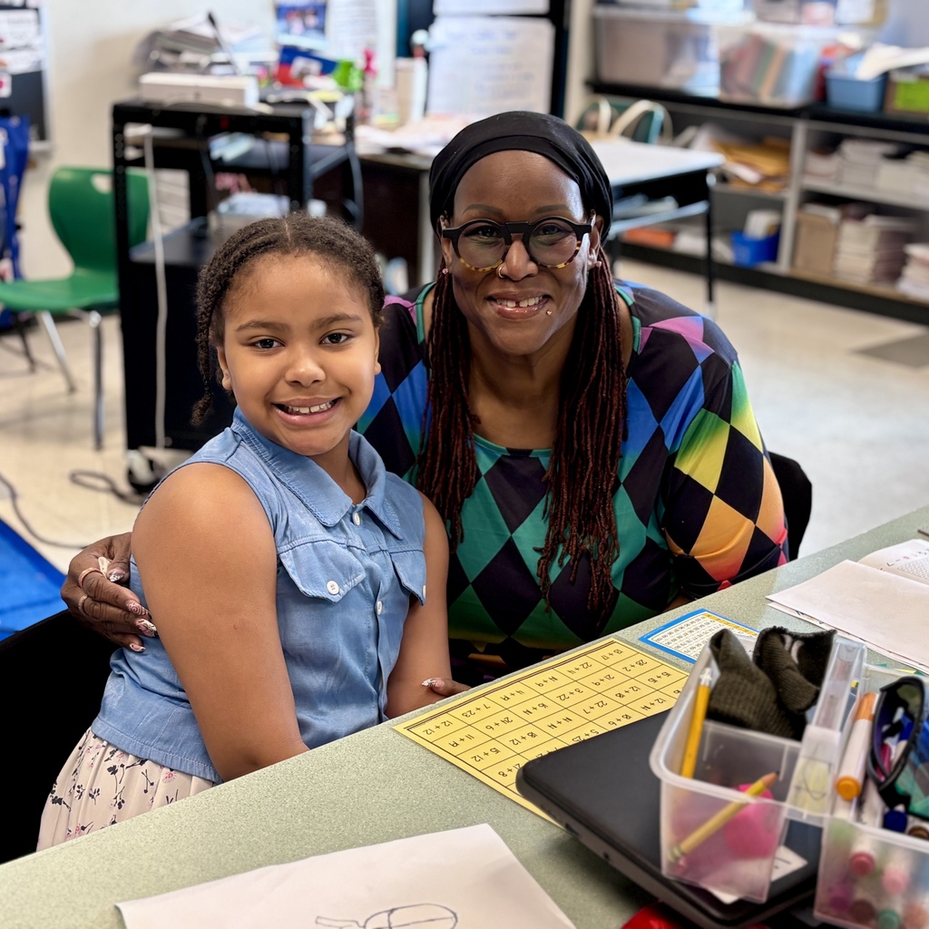 A smiling young girl and an adult woman pose together at a desk in a classroom.