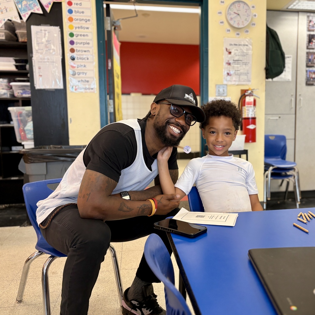 A man and a young boy smile at the camera while sitting at a blue table in a classroom.