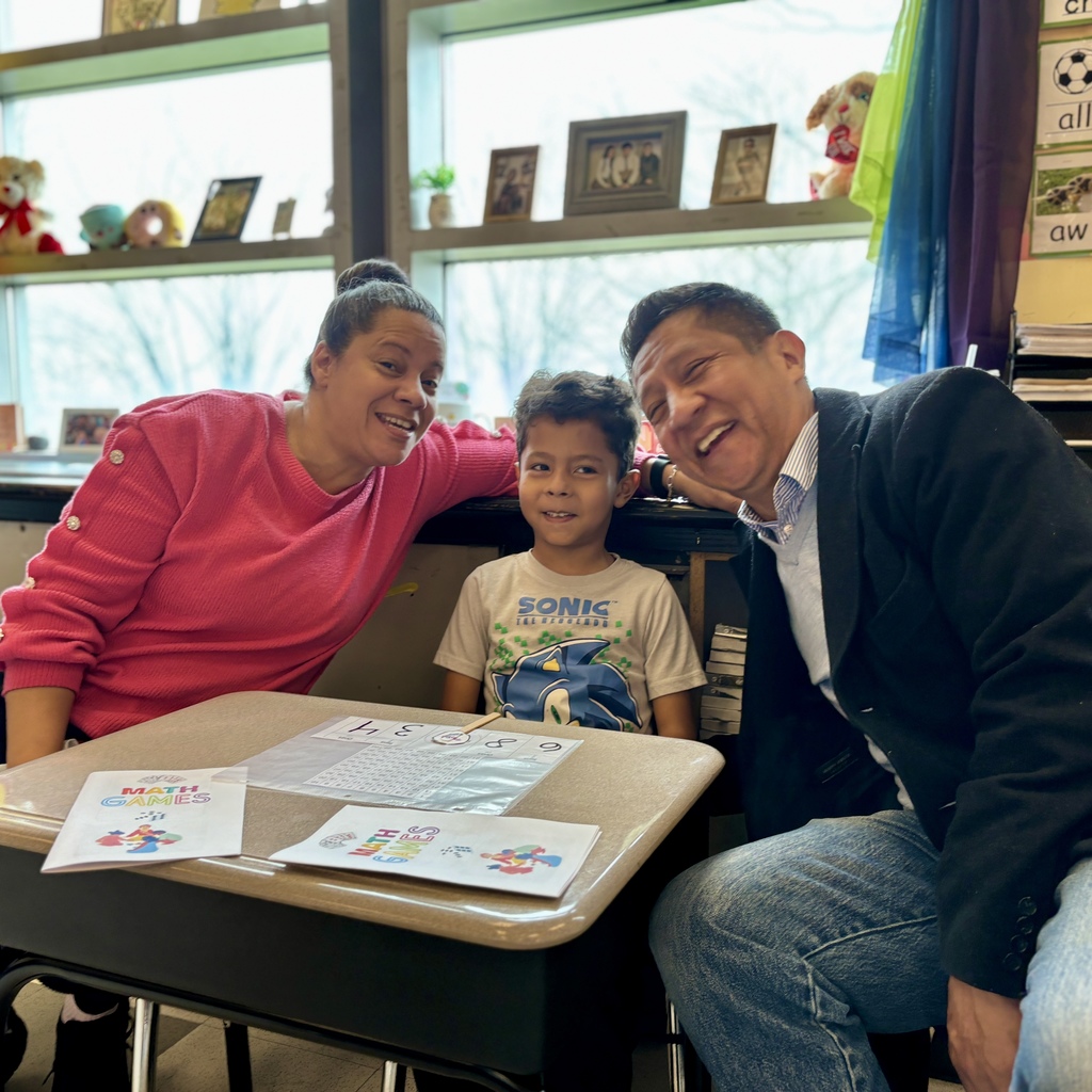 A smiling family poses for a photo at a desk in a classroom, with math games visible.
