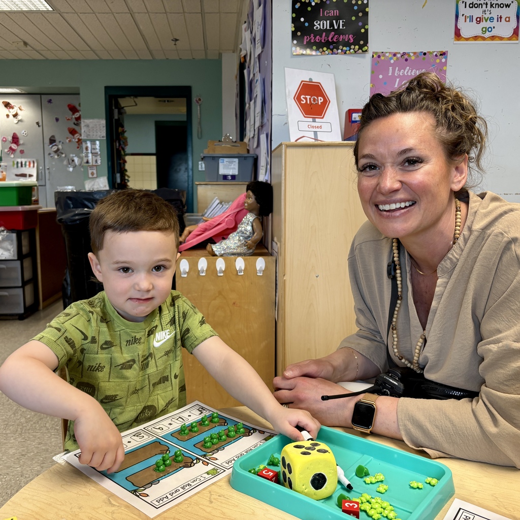A young boy and a woman smile while working on a math activity with frog counters and dice in a classroom.