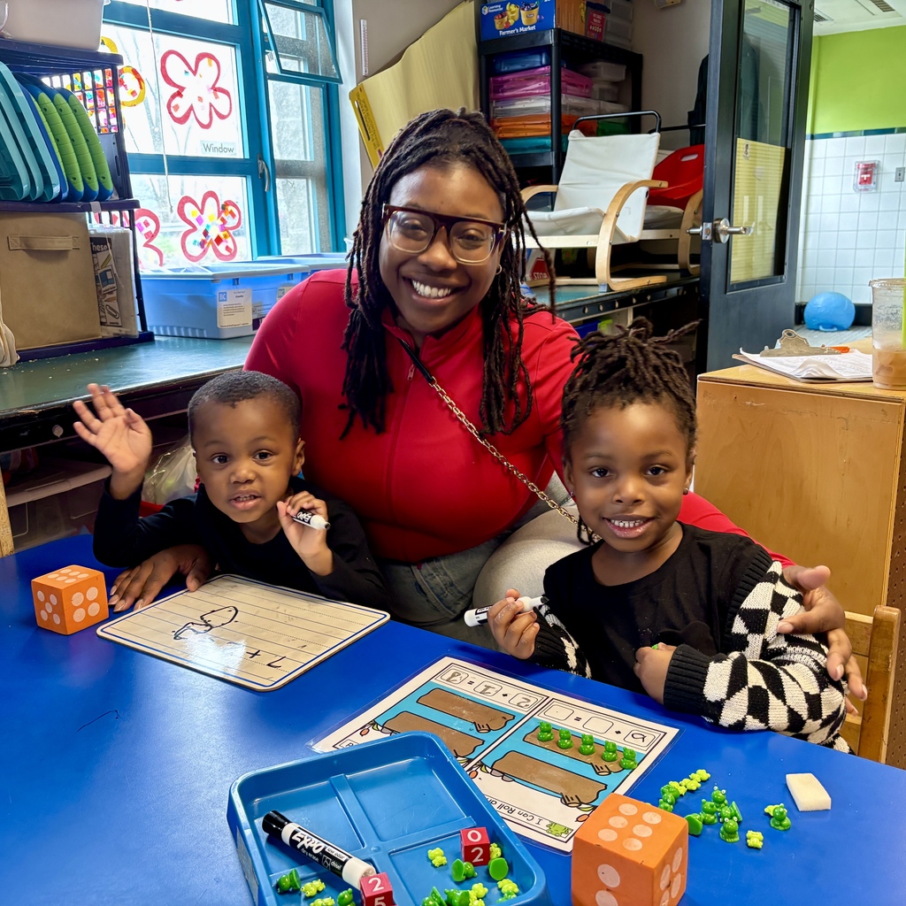 A teacher smiles with two young children at a blue table, engaged in a math lesson with dice and frog counters.