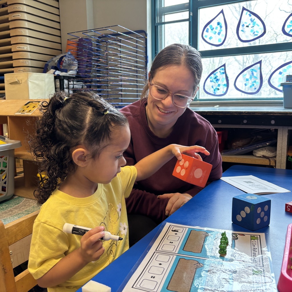 A teacher smiles while a young child plays with a large orange die and a marker at a blue table.
