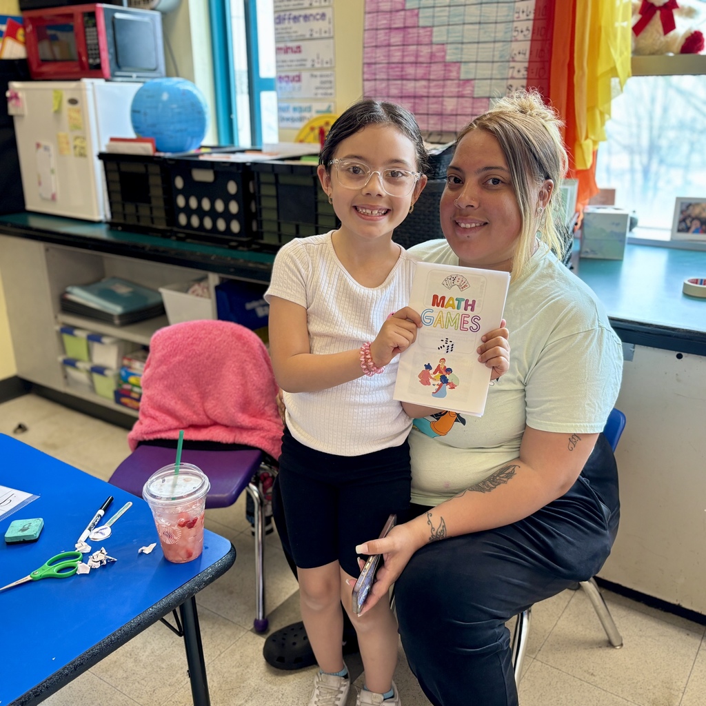 A young girl with glasses smiles while holding a booklet titled 'MATH GAMES' next to an adult.