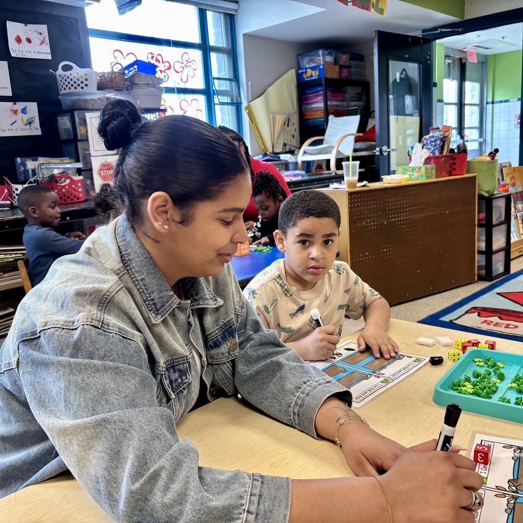 A teacher and children engage in a learning activity at a table in a classroom, with educational materials visible.
