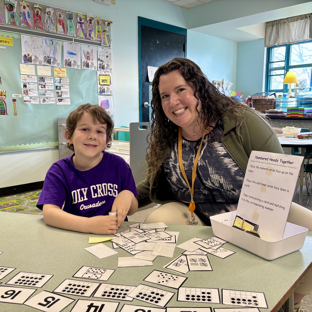 A smiling teacher and a young boy are engaged in a math game with number cards on a classroom table.