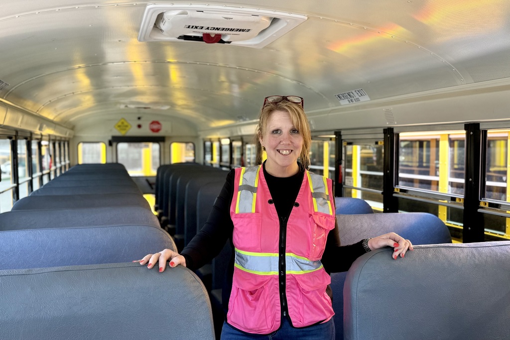 A woman in a pink safety vest stands inside a school bus, with rows of empty blue seats stretching behind her.