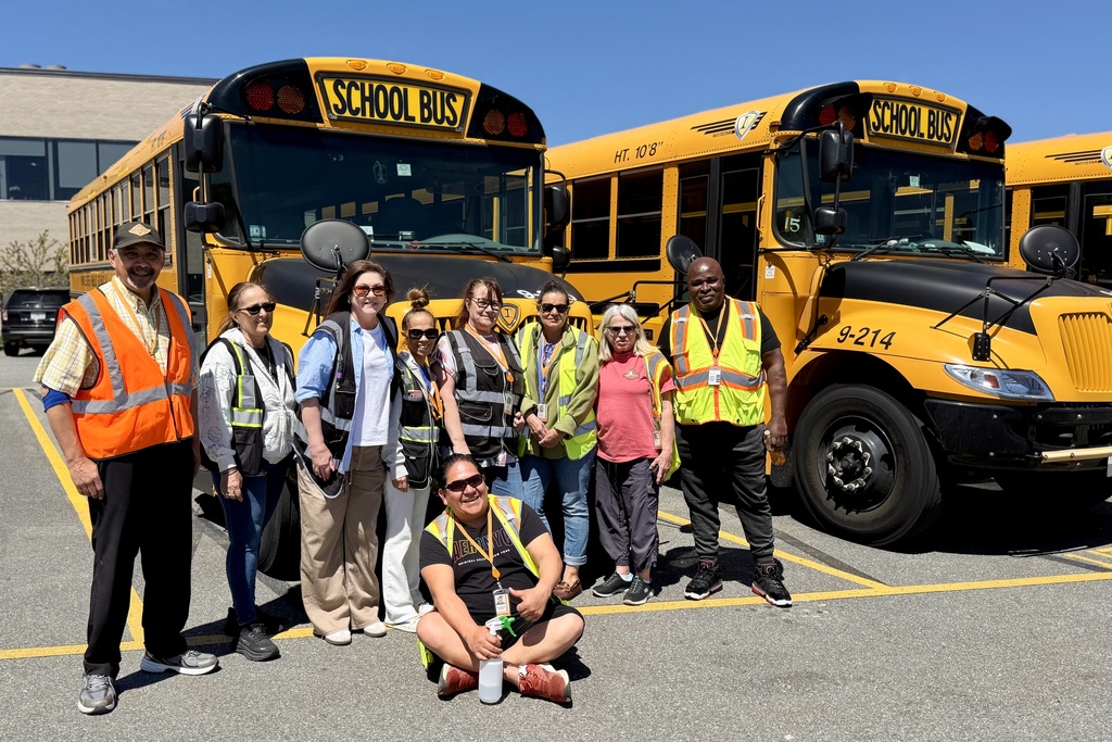 A group of school bus drivers and staff pose for a photo in front of yellow school buses.
