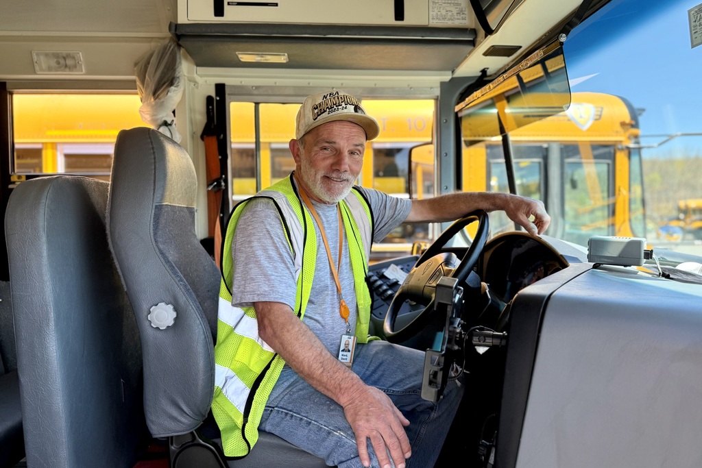 A smiling man wearing a reflective vest and a baseball cap sits in the driver's seat of a school bus.