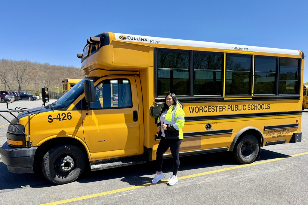 A woman in a high-visibility vest stands next to a yellow school bus labeled "WORCESTER PUBLIC SCHOOLS".