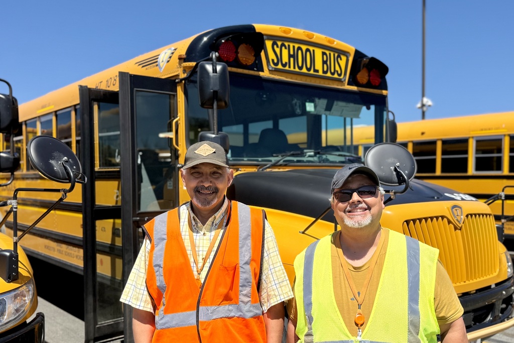 Two men in reflective vests stand in front of a yellow school bus.