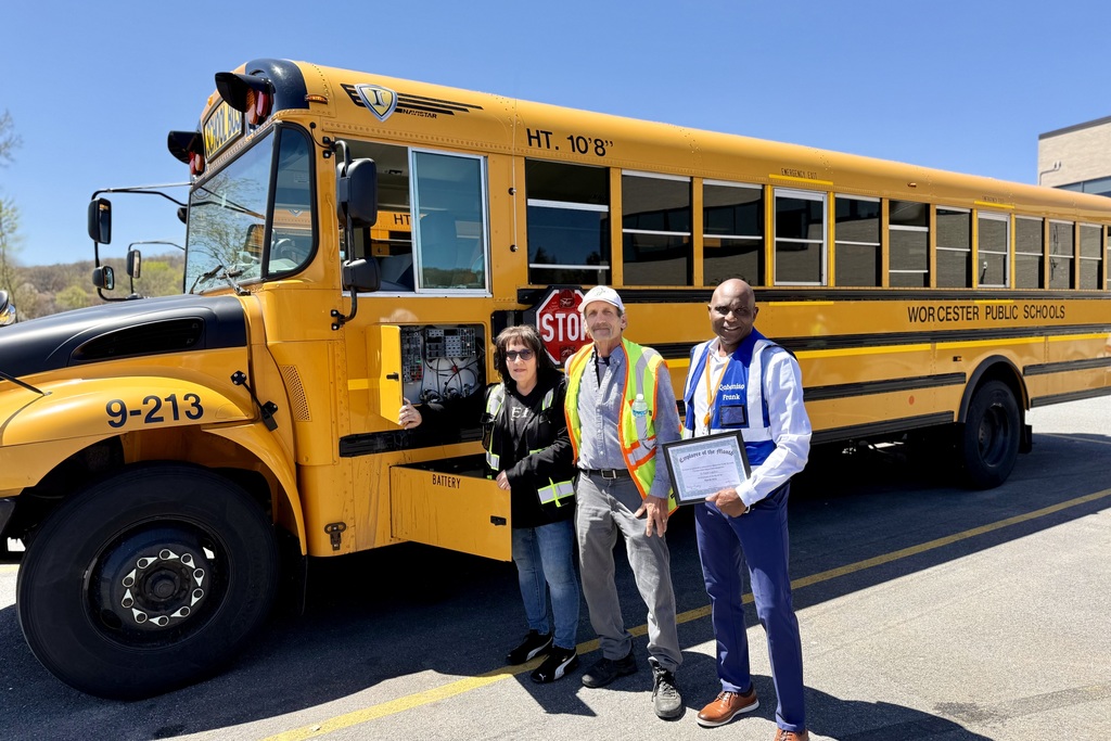 Three individuals stand in front of a yellow Worcester Public Schools bus, with one holding a certificate.