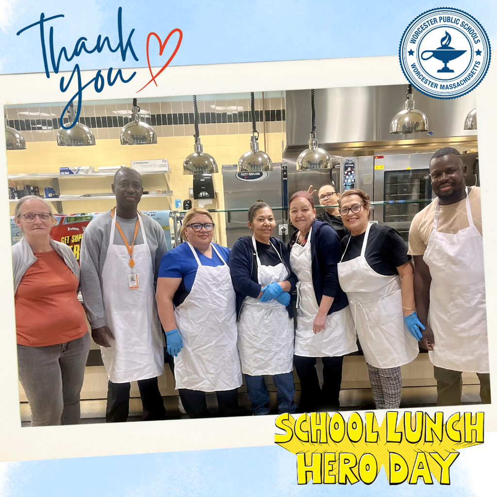 A group of smiling individuals in aprons stand together in a school cafeteria kitchen, celebrating School Lunch Hero Day.