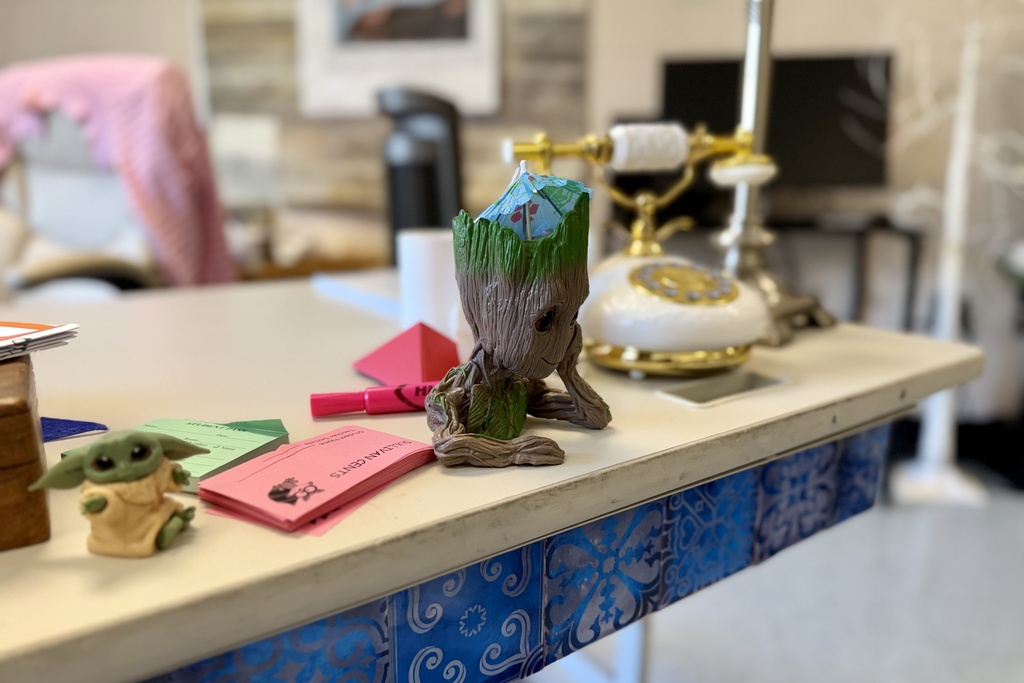 A small Groot figurine with a cocktail umbrella sits on a desk next to a Baby Yoda figure and pink and green slips of paper.