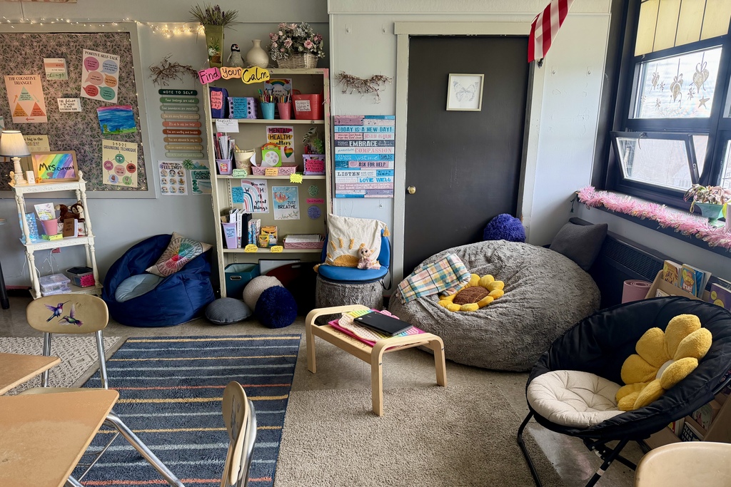 A cozy classroom corner features beanbag chairs, rugs, and a bookshelf filled with books and art supplies.