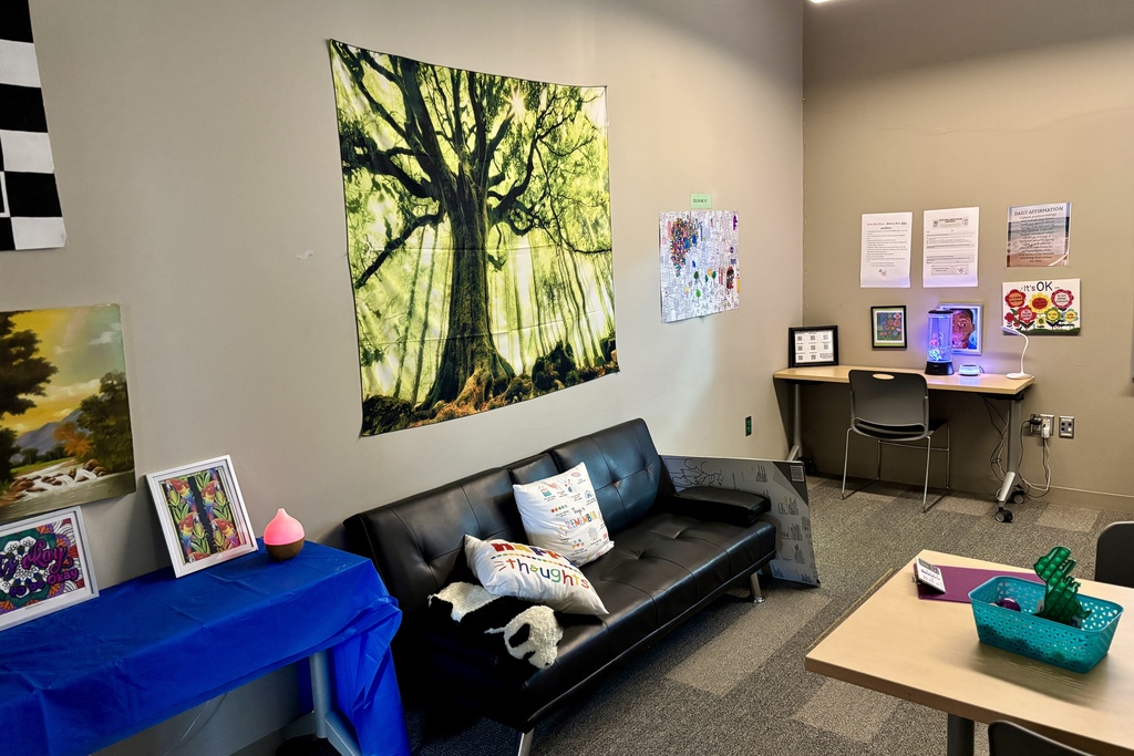 A calming room features a black leather couch with decorative pillows, a large tapestry of a sunlit forest, and a desk with a lava lamp.