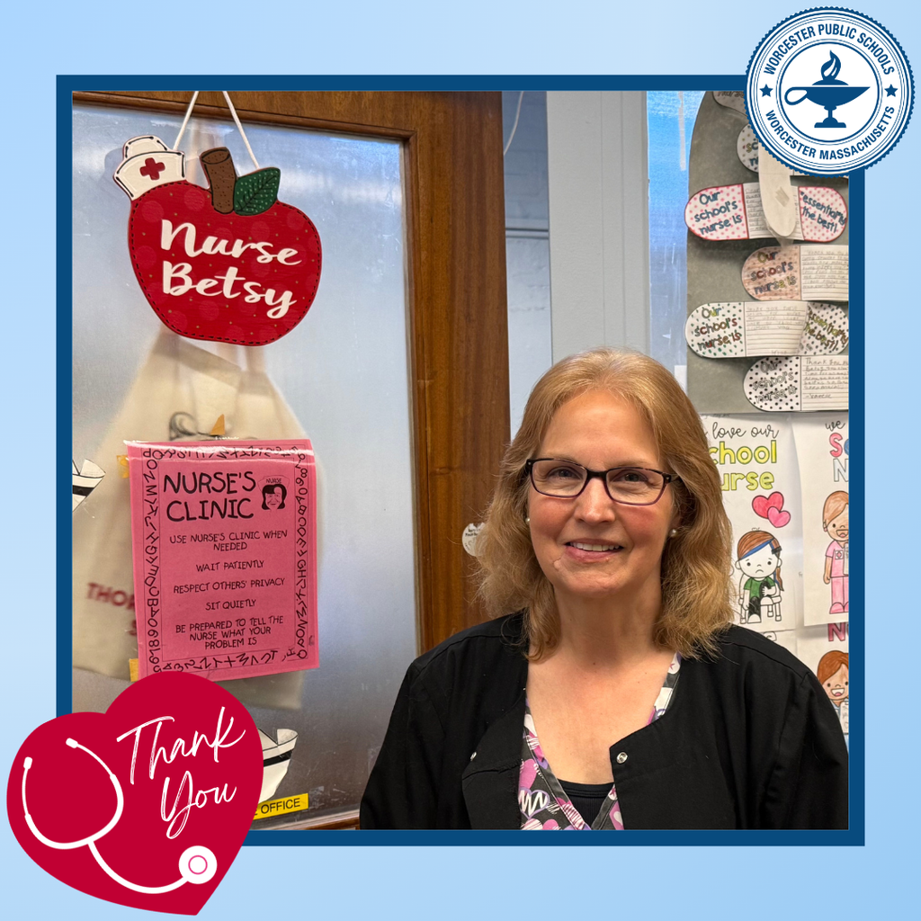 A smiling school nurse stands in front of a door decorated with an apple sign and a 'Nurse's Clinic' notice.