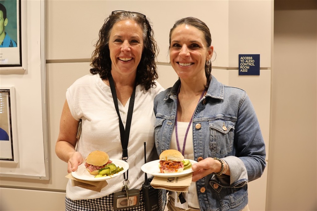 Two smiling women hold plates with sandwiches and pickles, standing indoors.
