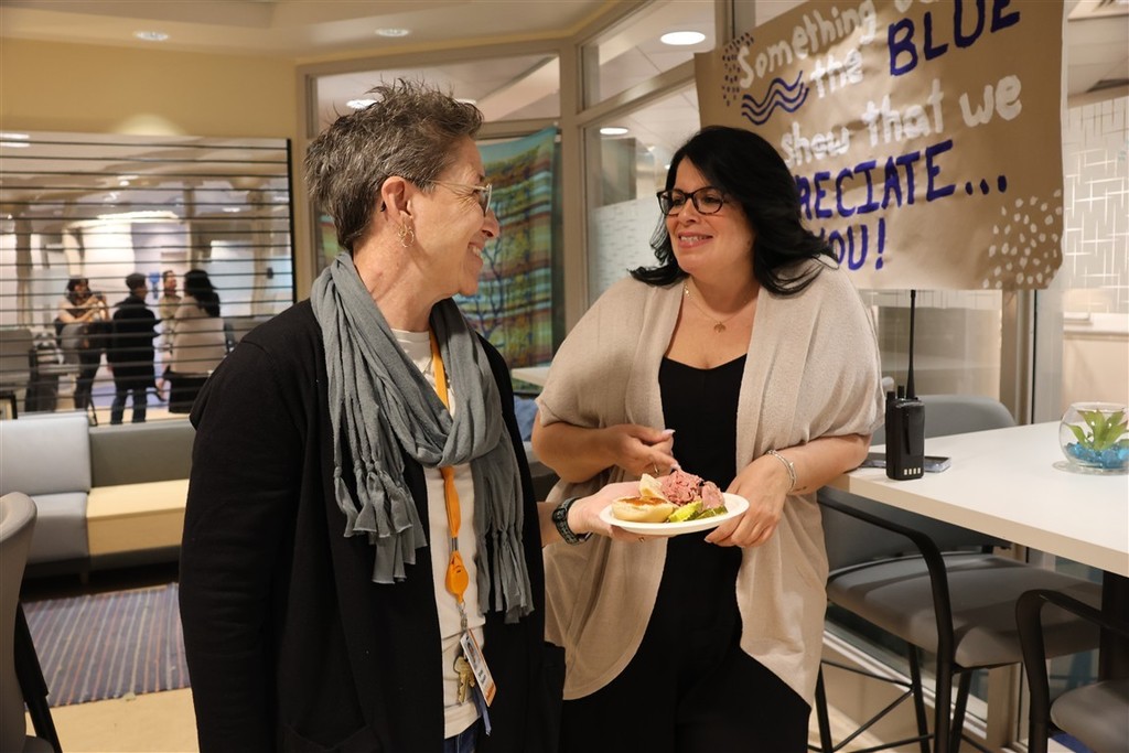 Two women smile and chat while one holds a plate with a sandwich and pickles.