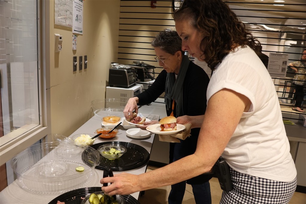 Two women prepare sandwiches at a buffet table with various toppings.