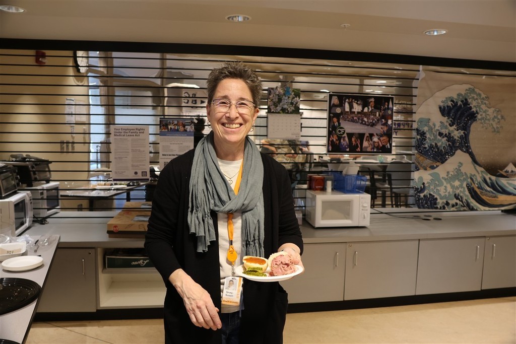 A smiling woman holds a plate of food, including a sandwich and pickles, in a breakroom.