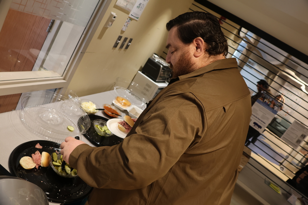 A man in a brown jacket prepares a sandwich with pickles and meat at a buffet.