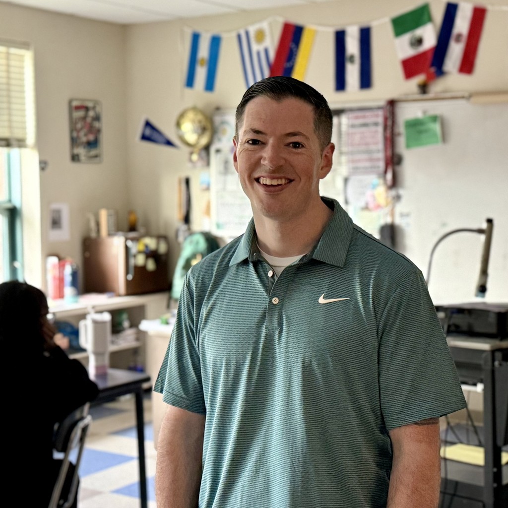 A smiling man in a striped polo shirt stands in a classroom decorated with international flags.