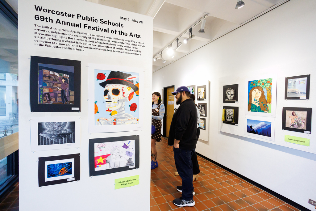 Patrons stand in a hallway observing artwork while text is visible on the wall that says "Worcester Public Schools 69th Annual Festival of the Arts"