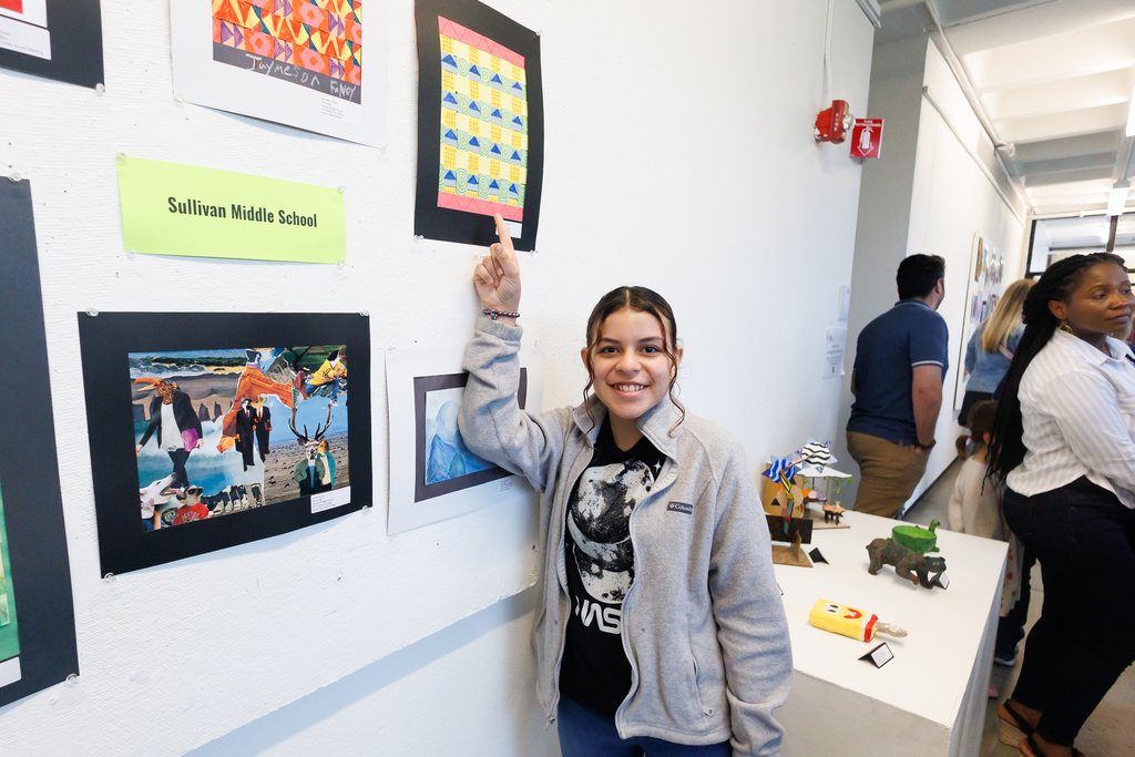 A student points up toward their artwork hanging on a wall in a gallery.