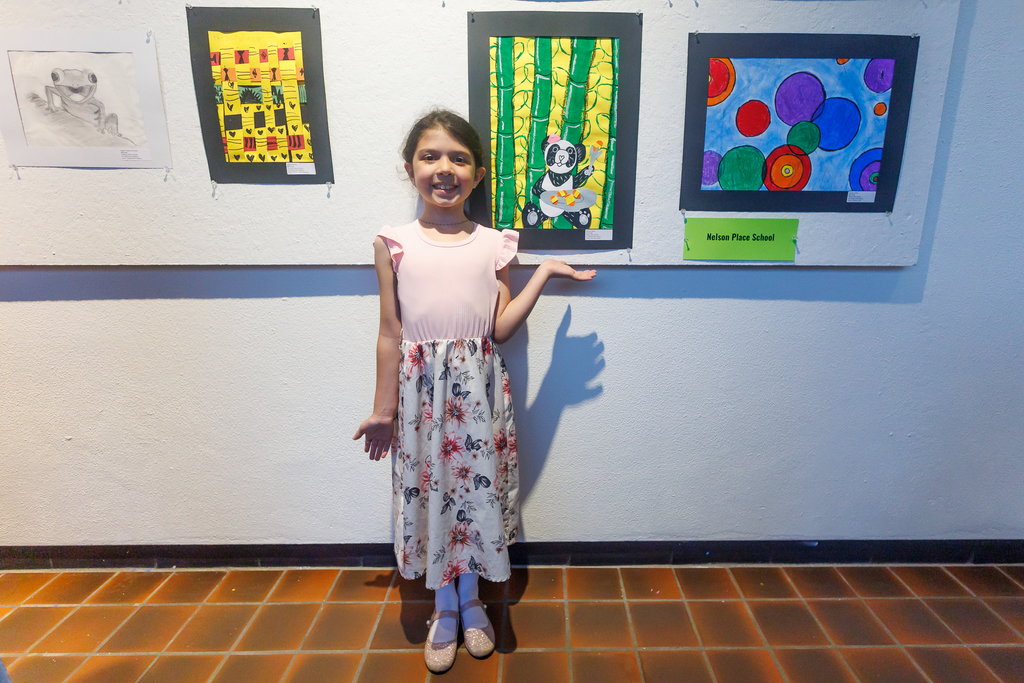 A student holds out their hand under their artwork, which is displayed on a wall alongside other paintings. 