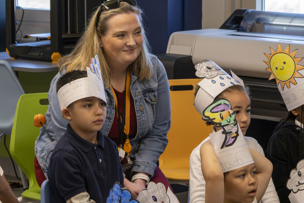 A teacher smiles while observing children wearing handmade paper crowns decorated with weather symbols.