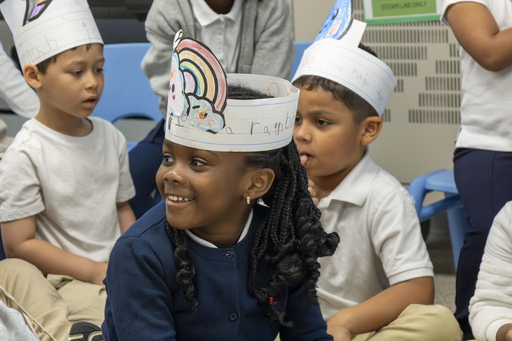 Children wearing handmade paper crowns with drawings of rainbows and faces sit together.