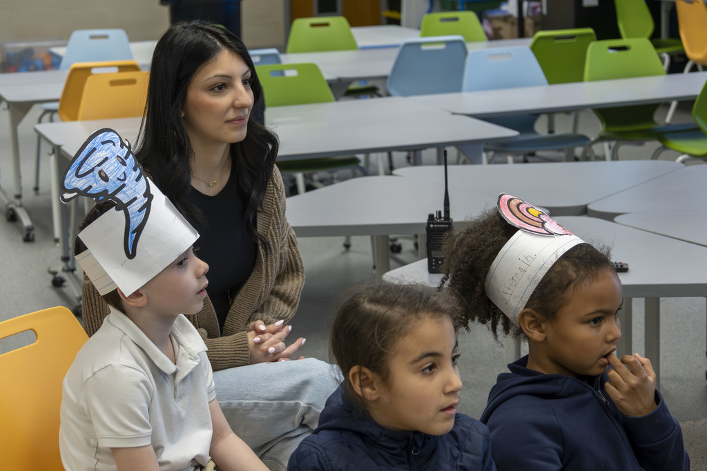 A teacher and three young students wearing paper hats sit at desks in a classroom