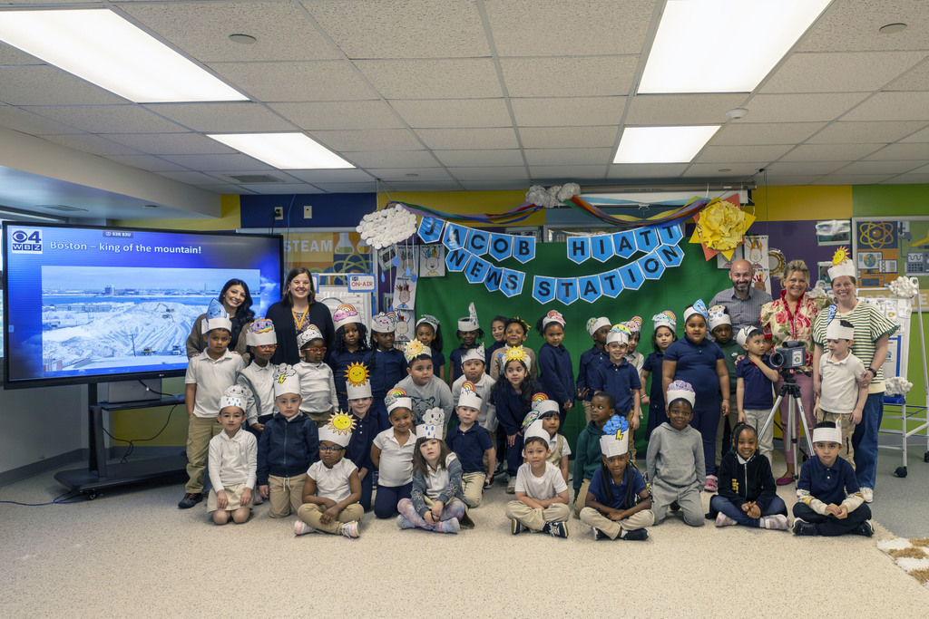 A group of children and adults pose for a photo in a classroom decorated with a "Jacob Hiatt News Station" banner.
