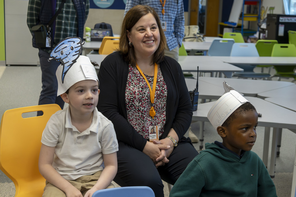 A smiling woman in a patterned shirt and black cardigan sits with two young boys wearing paper hats in a classroom.