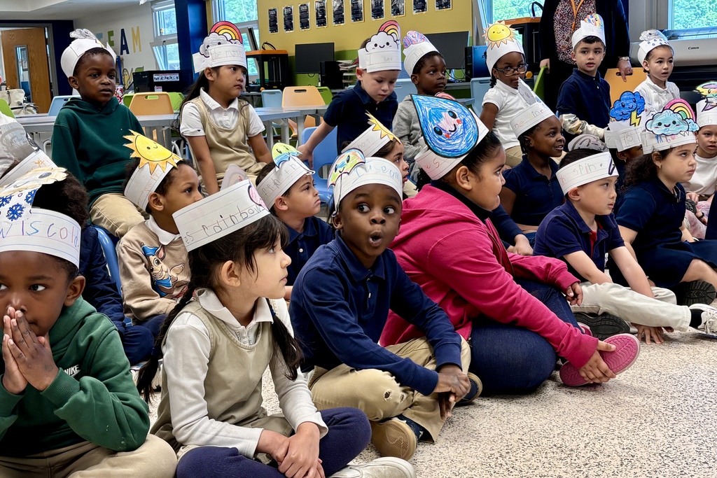 A diverse group of young students wearing handmade paper hats sit on the floor in a classroom.