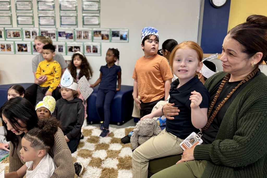 Children and adults gather in a classroom, some wearing paper hats, with artwork displayed on the wall.