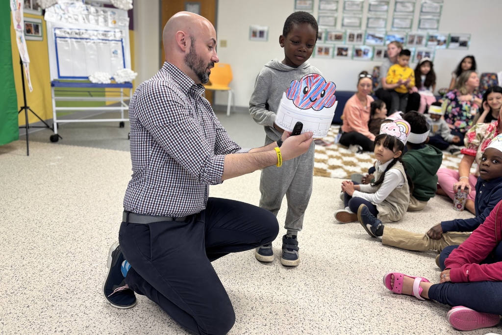 A teacher kneels to show a child a craft project in a classroom with other children sitting on the floor.