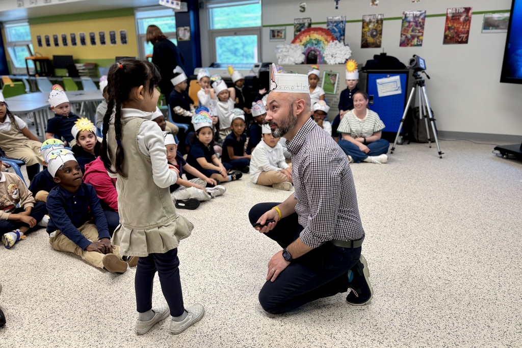 A teacher kneels to talk with a young student in a classroom filled with children wearing festive paper hats.