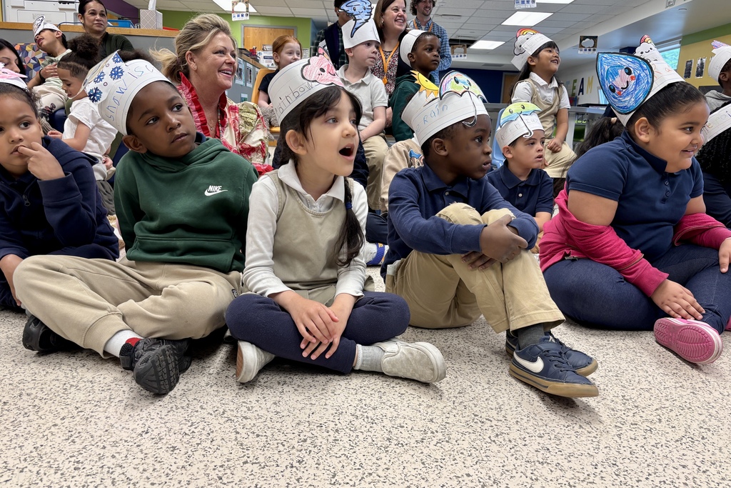 Children in a classroom wear festive paper hats decorated with weather symbols and drawings.