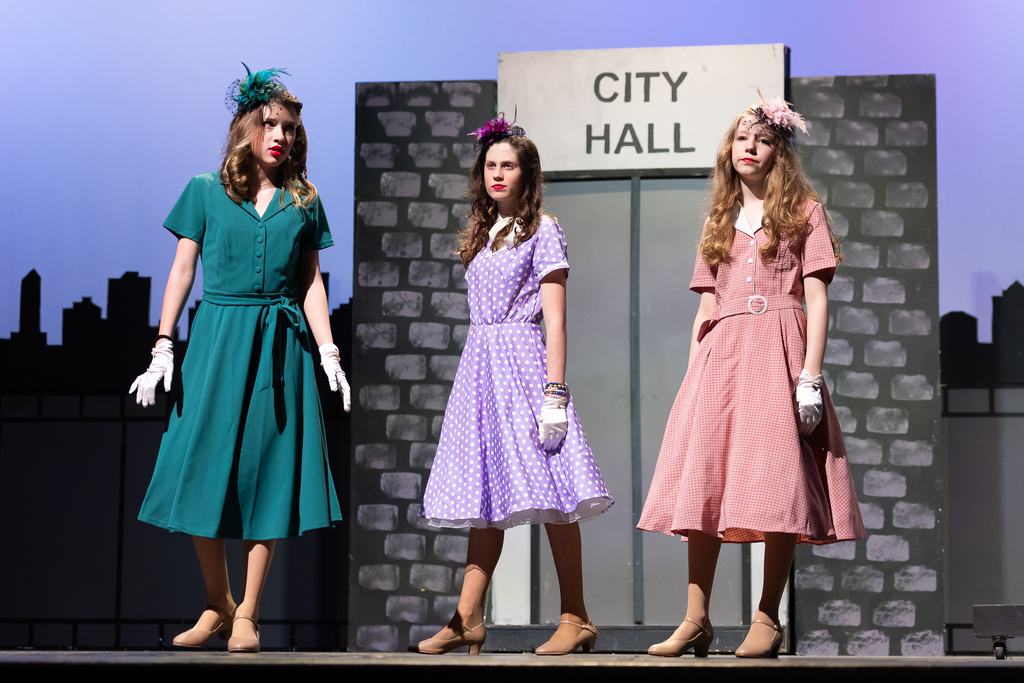 Three students wearing colorful dresses perform on stage during a play.