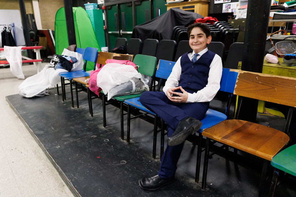 A student dressed in a costume sits in a chair and smiles while behind the scenes.
