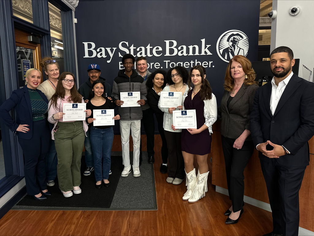 A group of students and adults stand together in front of a Bay State Bank sign, holding scholarship certificates.
