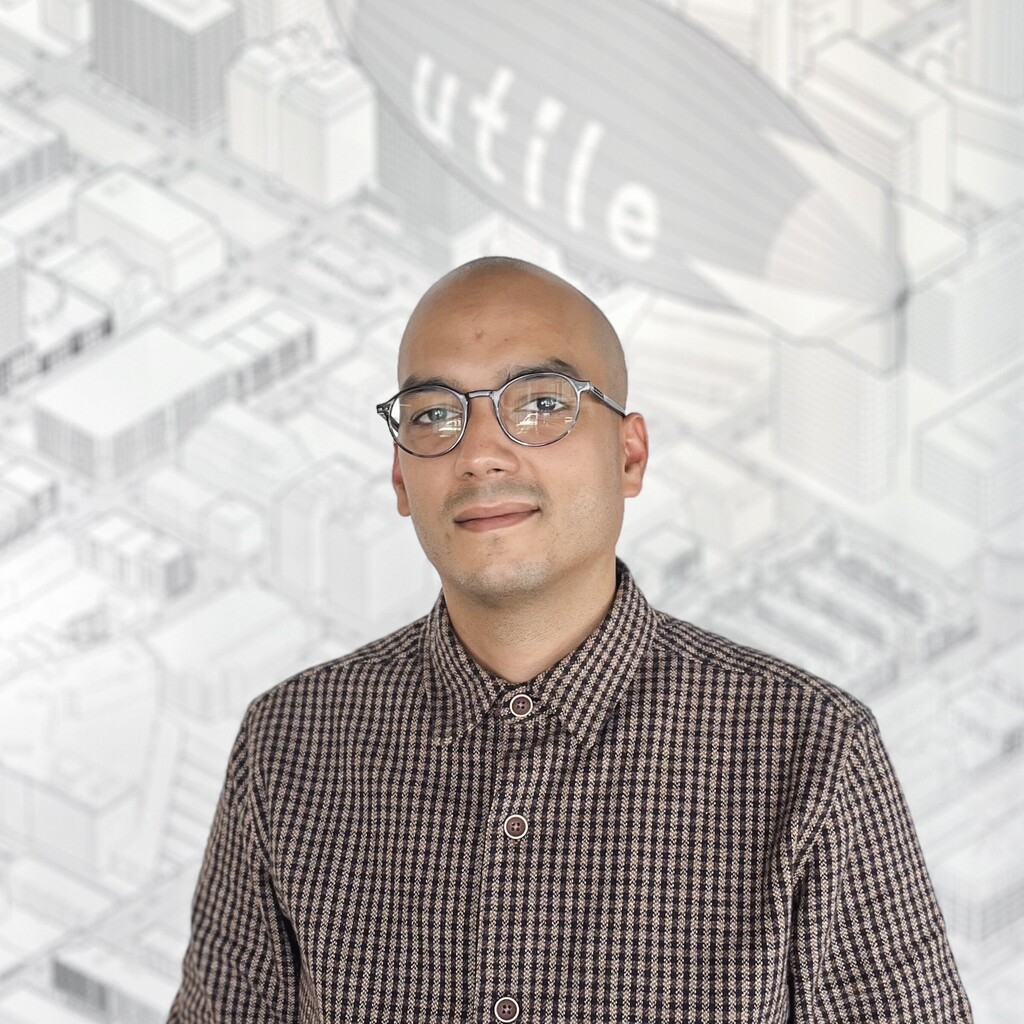 A man with a shaved head and glasses smiles, wearing a checkered shirt against a blueprint background.