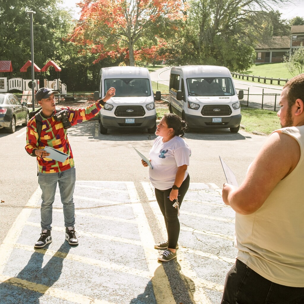 Three people stand in a parking lot, with one person gesturing and holding papers.