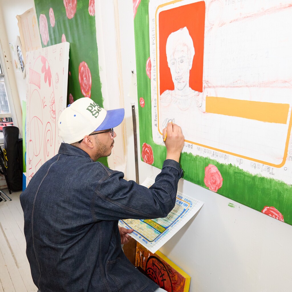An artist in a denim jacket and a baseball cap sketches on a large canvas featuring a portrait and floral elements.