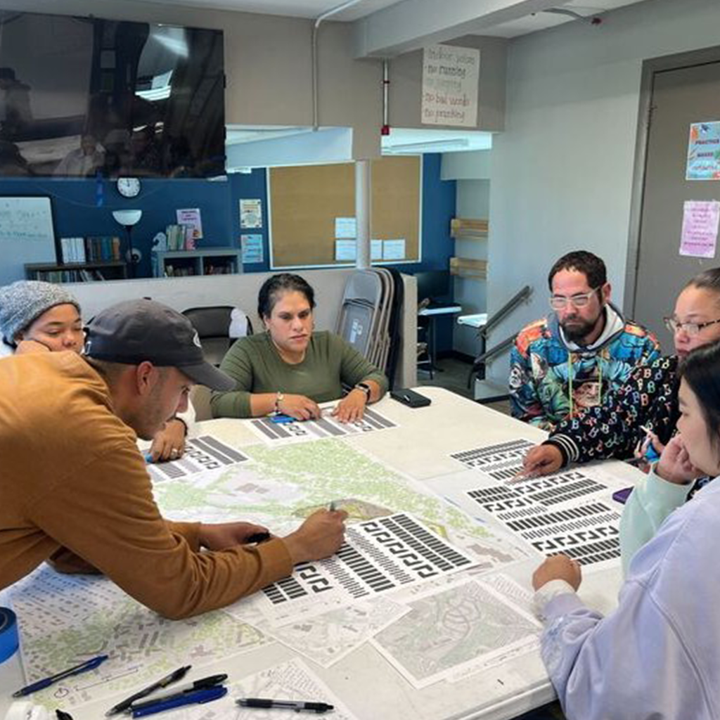 A diverse group of people collaborates around a table covered with maps and blueprints.