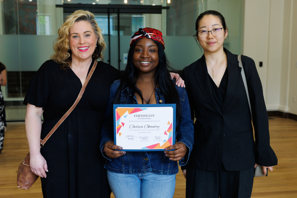 A high school senior stands between two teachers while holding their award certificate.
