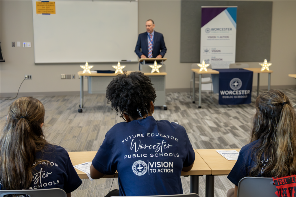 Three students in navy blue shirts with 'Worcester Public Schools Future Educator' text sit at desks facing a speaker.