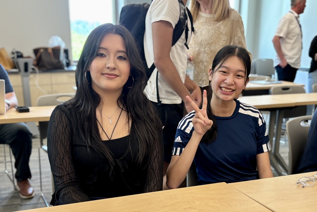 Two young women smile at the camera while seated at a table in a classroom or meeting room.